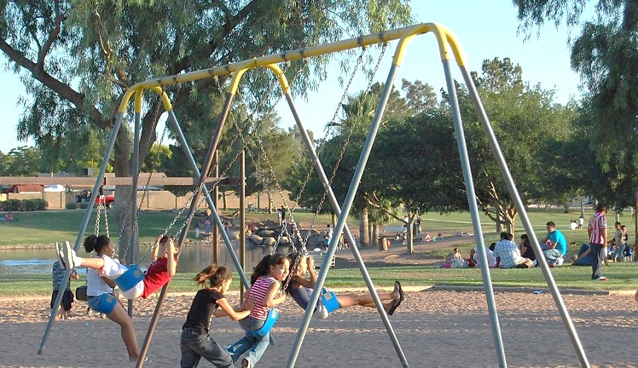 Children play on a swing set in the foreground. In the background, children and families play in the park.